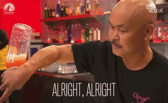 a man in a black shirt is holding a glass and says alright
