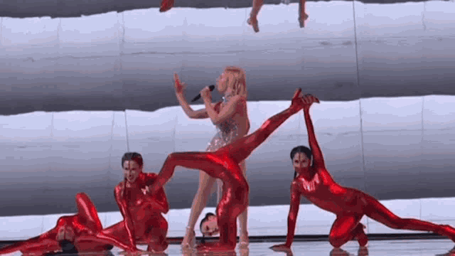 a woman singing into a microphone while a group of red dancers perform