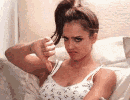 a woman in a white tank top is sitting on a bed giving a thumbs down sign .