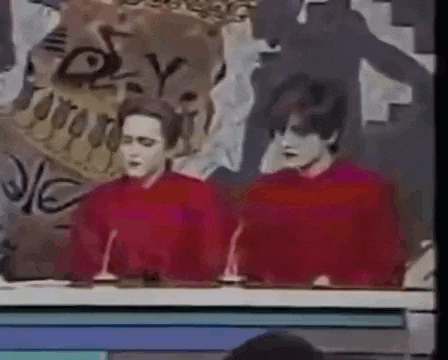 two men in red sweaters are sitting at a table with microphones in front of a wall with graffiti on it .