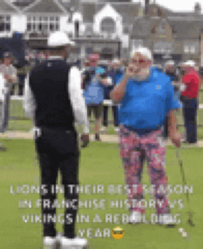 a man in a blue shirt is standing on a golf course talking to another man .