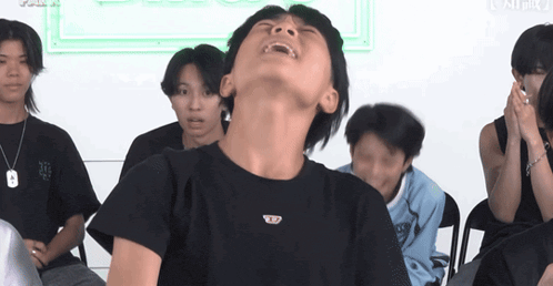 a group of young men are sitting in a room with one wearing a black t-shirt with the letter b on it