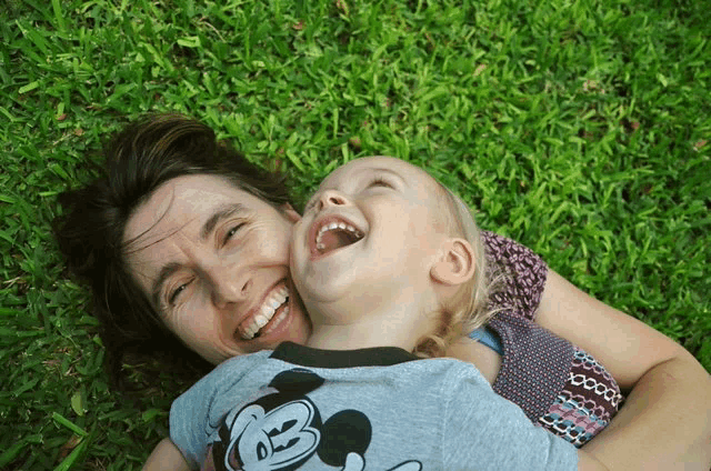 a woman and child are laying in the grass and the child has a mickey mouse shirt on