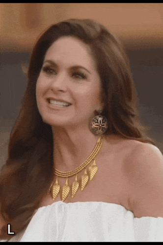 a woman wearing a white off the shoulder top and gold necklace smiles