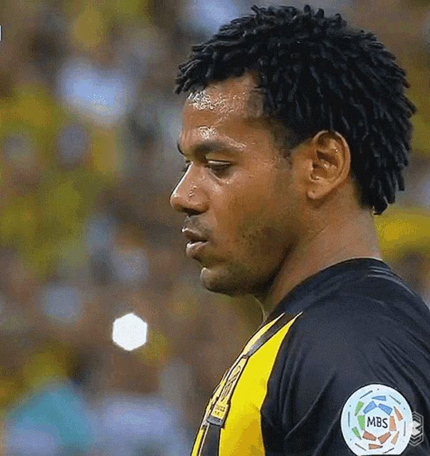 a man with dreadlocks is wearing a black and yellow shirt with a mbs patch on his arm