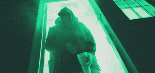 a person in a hooded jacket stands in a doorway with a green light behind them