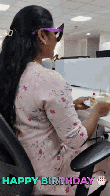 a woman wearing sunglasses sits at a desk holding a piece of cake with the words happy birthday written on the bottom