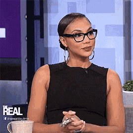 a woman wearing glasses and a black dress is sitting at a table with her hands folded and a cup of coffee .