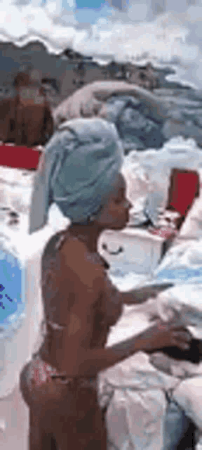 a woman in a bikini with a towel wrapped around her head is standing next to a pile of blankets .