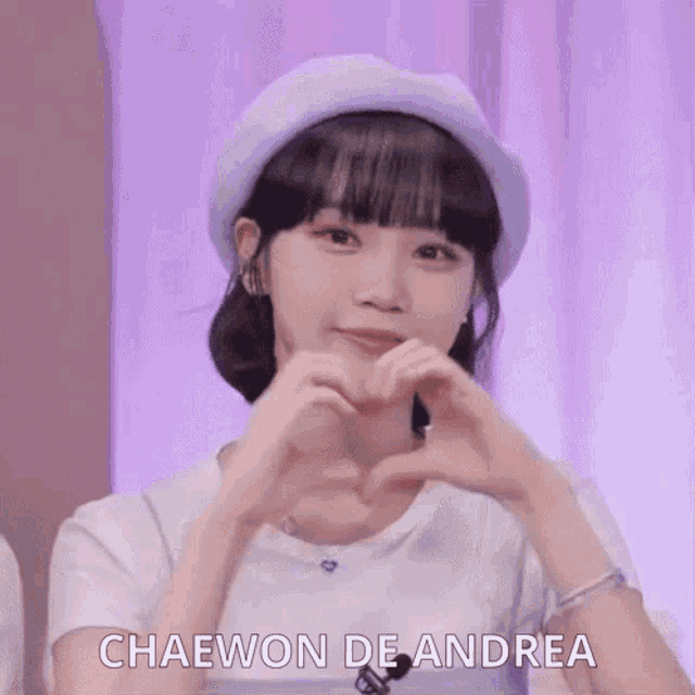 the girl is wearing a purple hat and making a heart with her hands .