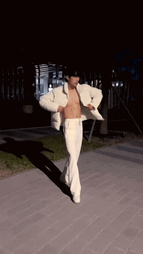 a shirtless man in a white jacket and white pants is walking on a sidewalk at night