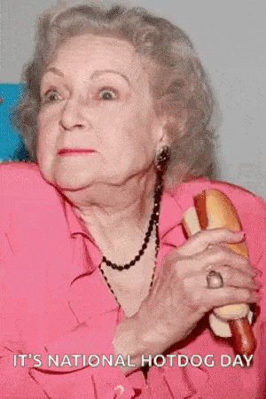 an elderly woman in a pink shirt is holding a hot dog and says it 's national hotdog day .