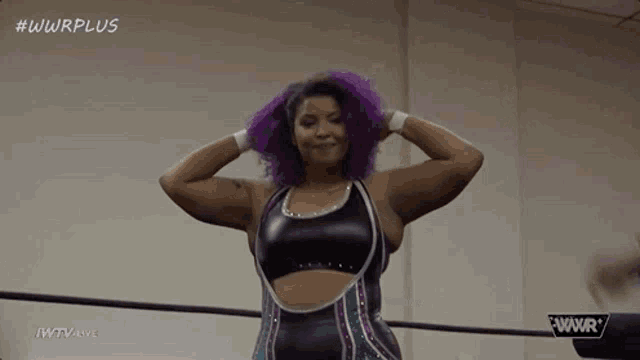 a woman with purple hair is standing in a wrestling ring with her hands behind her head .