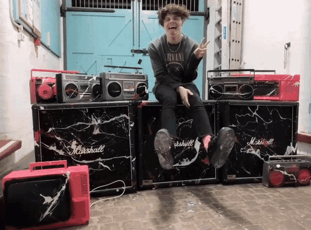 a man wearing a nirvana shirt is sitting on a marshall amplifier
