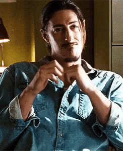 a man with a beard wearing a denim shirt is sitting in a chair with his hands folded .