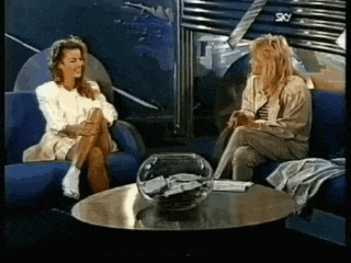 two women are sitting on a blue couch talking to each other in front of a sky sign