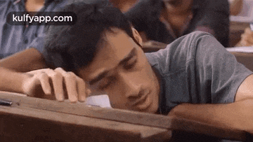 a young man is sleeping on a wooden desk in a classroom .