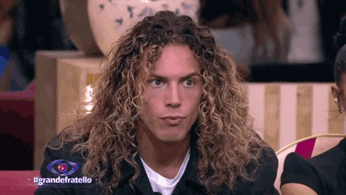 a man with long curly hair looks at the camera in front of a sign that says grandefratello