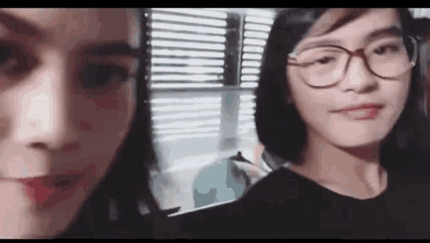 two women are standing next to each other in front of a window . one of the women is wearing glasses .
