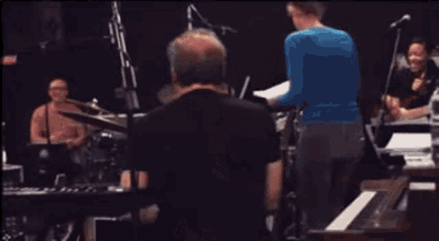a man in a black shirt is sitting at a piano with a woman playing drums in the background