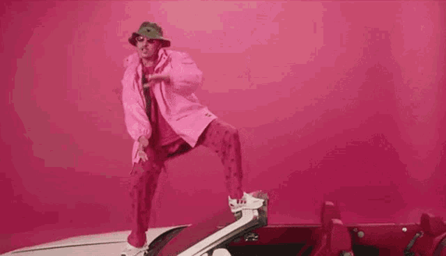 a man in a pink jacket and hat is standing on top of a pink convertible car .