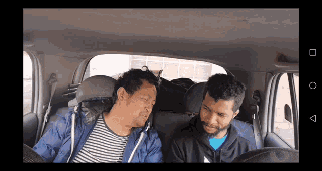 two men are sitting in the back seat of a car and one is wearing a blue jacket