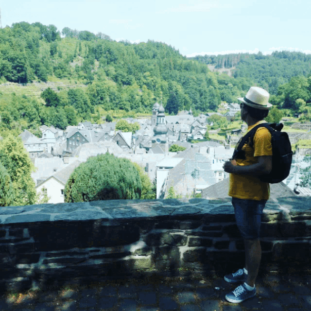 a man with a backpack stands in front of a stone wall overlooking a small town