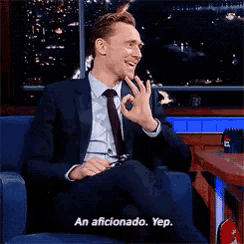 a man in a suit and tie is sitting in a chair and saying an aficionado yep
