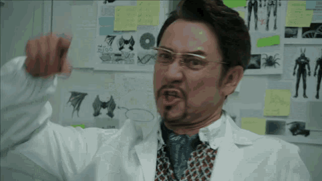 a man wearing glasses and a lab coat points to a wall with sticky notes on it