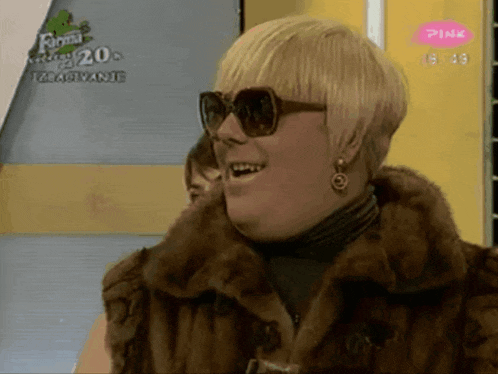 a woman wearing sunglasses and a fur coat is smiling in front of a pink sign