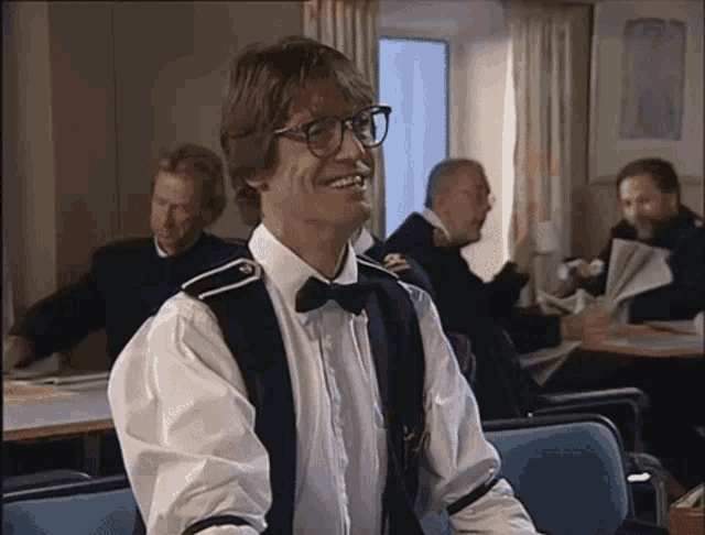 a man wearing glasses and a bow tie smiles in a room with other men