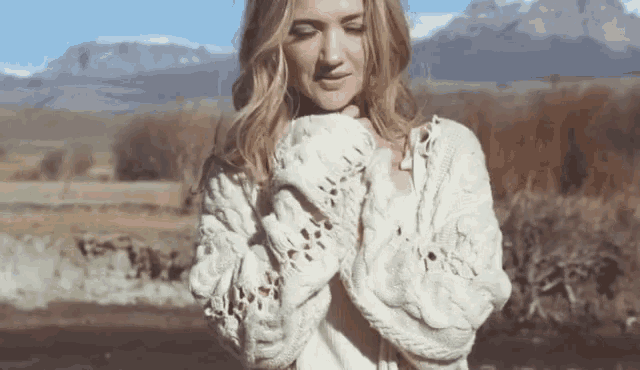 a woman wearing a white sweater and mittens is standing in front of a mountain .