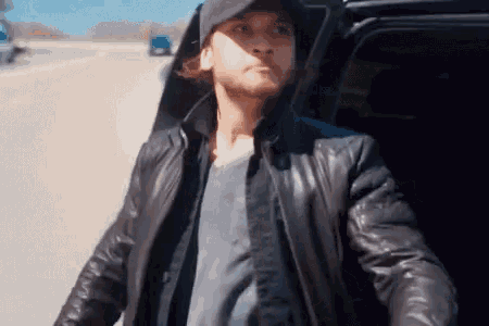 a man wearing a hat and a leather jacket is getting out of a car on a highway .