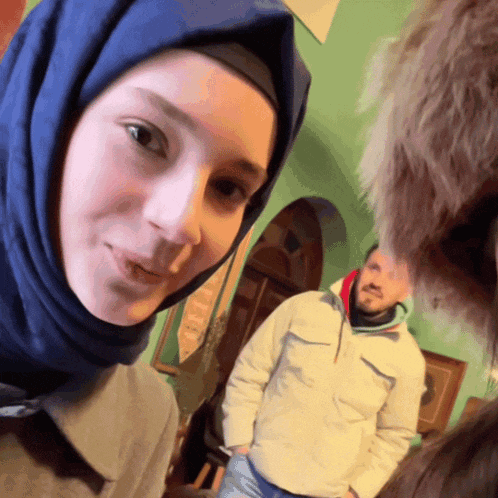 a woman wearing a blue hijab looks at the camera while a man in a white jacket stands behind her