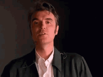 a man in a green jacket and white shirt is making a funny face in a dark room .