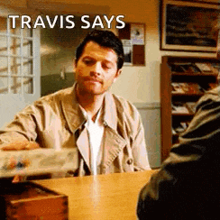 a man in a trench coat is sitting at a table with a box of money in front of him .