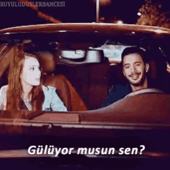 a man and a woman are sitting in a car and the man is smiling and the woman is asking " gülüyor musun sen "