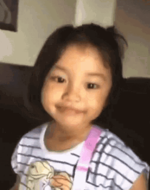 a little girl is smiling and looking at the camera while wearing a striped shirt and a pink backpack .
