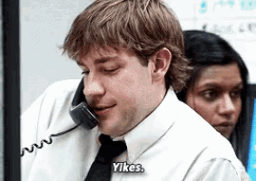 a man in a white shirt and black tie is talking on a phone and says " yikes "