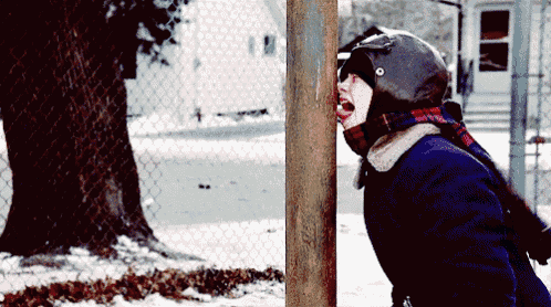 a young boy wearing a helmet and scarf is sticking his head through a chain link fence in the snow