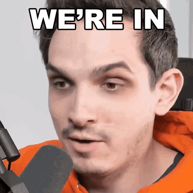 a man in an orange hoodie is talking into a microphone and says we 're in