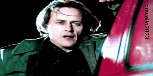 a man in a green coat is standing next to a red car with the year 2003 written on the bottom