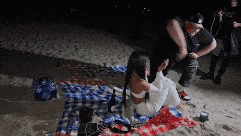a woman sits on a plaid blanket on the beach with a man standing behind her