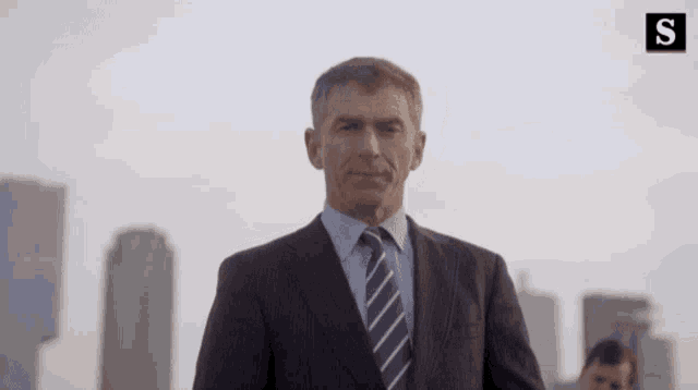 a man in a suit and tie stands in front of a city skyline with the letter s on the bottom