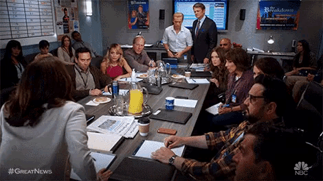 a group of people are sitting around a table in a conference room with a nbc logo on the wall .