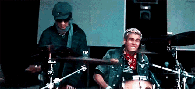 two men are playing drums on a stage and one of them has a bandana around his neck .