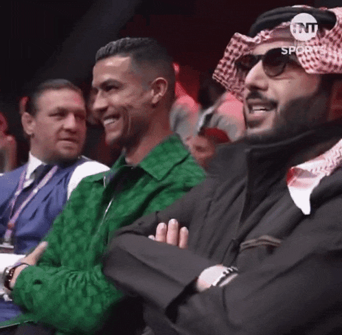 a group of men are sitting in a boxing ring and one of them is wearing a turban .