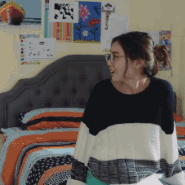 a woman wearing glasses and a striped sweater stands in front of a bed