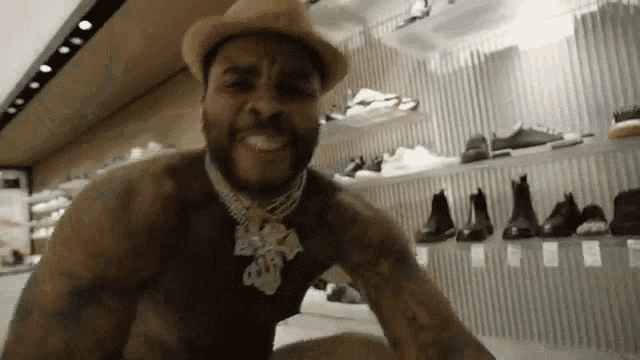 a shirtless man wearing a hat and a necklace is standing in front of a wall of shoes .