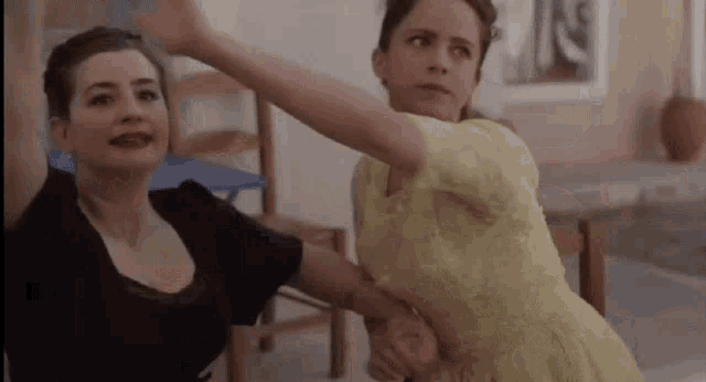 two women are standing next to each other in a room . one of the women is wearing a yellow dress .